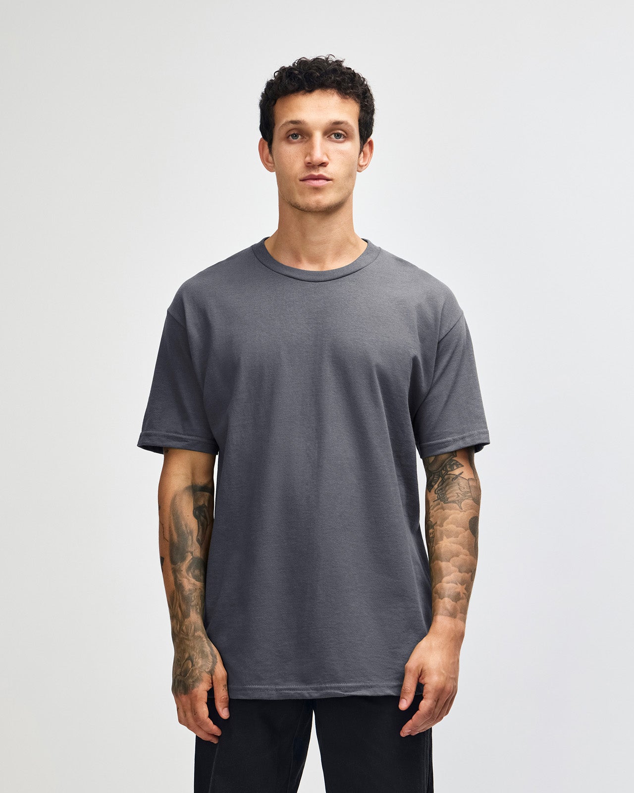 Male Model wearing a Charcoal Heavyweight Unisex Short Sleeve Crew Neck Tee, paired with blue jeans, standing against a plain background (front pose) -charcoal