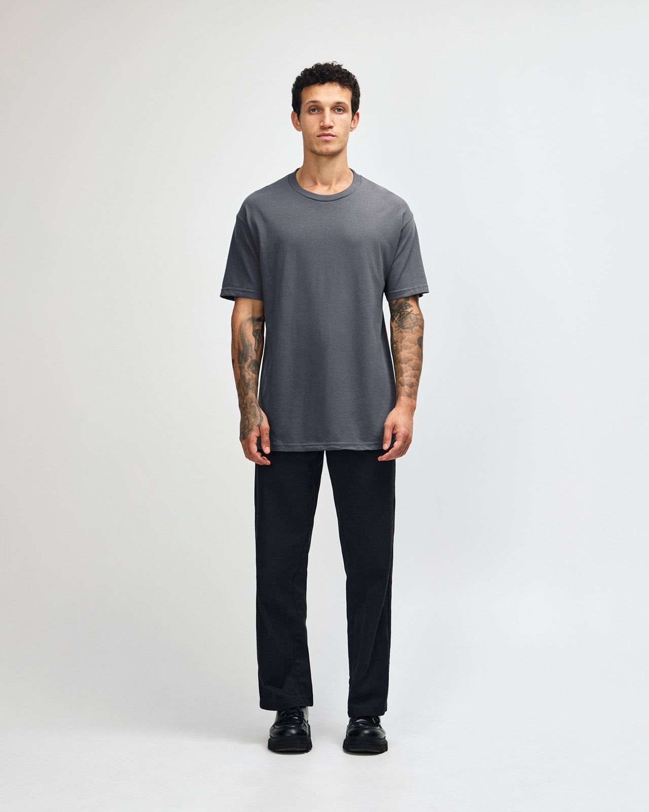 Male Model wearing a Charcoal Heavyweight Unisex Short Sleeve Crew Neck Tee, paired with blue jeans, standing against a plain background (front pose) -charcoal