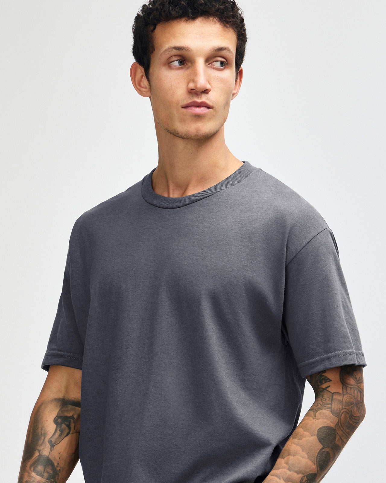 Male Model wearing a Charcoal Heavyweight Unisex Short Sleeve Crew Neck Tee, paired with blue jeans, standing against a plain background (front pose) -charcoal