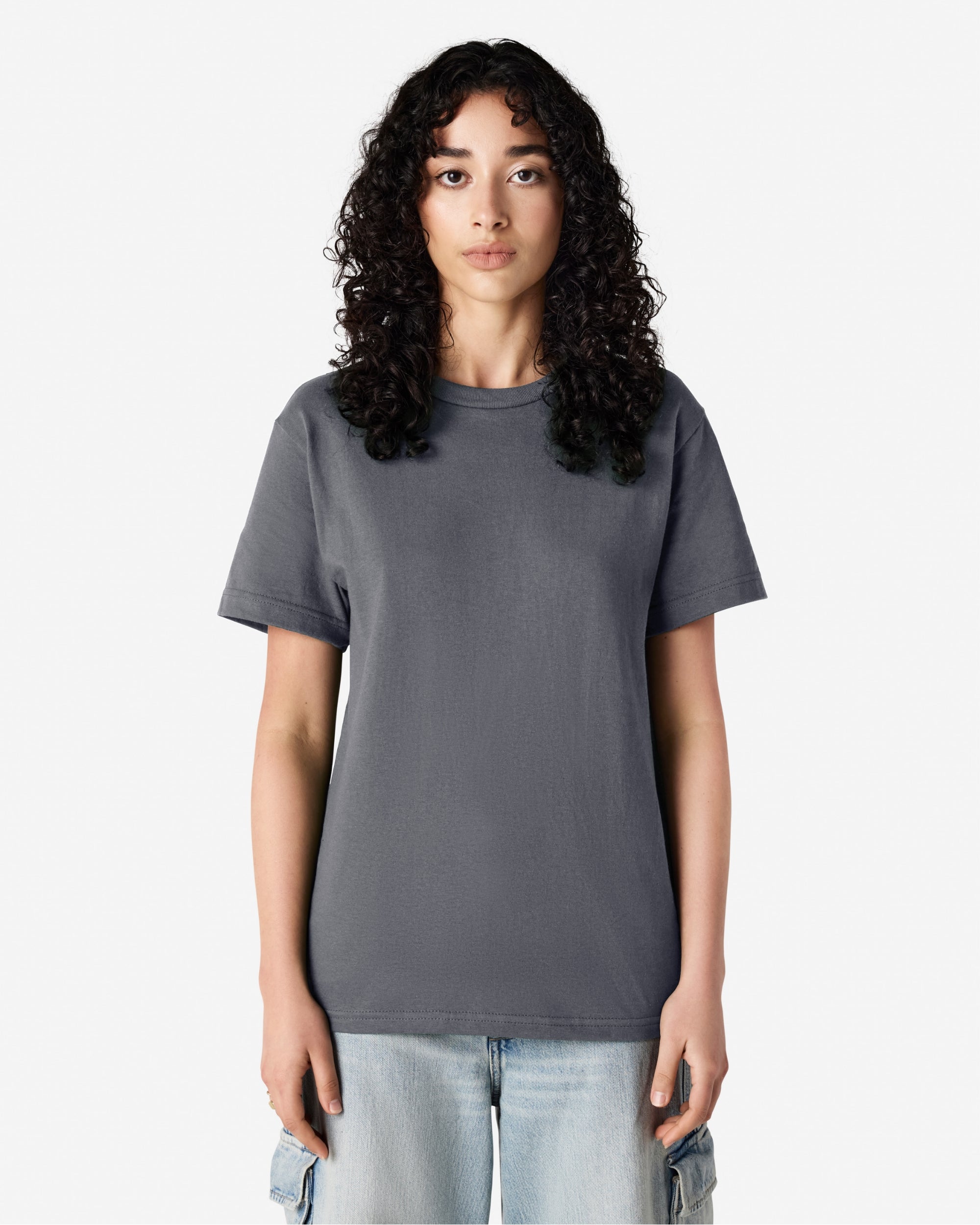 Female Model wearing a Charcoal Heavyweight Unisex Short Sleeve Crew Neck Tee, paired with blue jeans, standing against a plain background (front pose) -charcoal