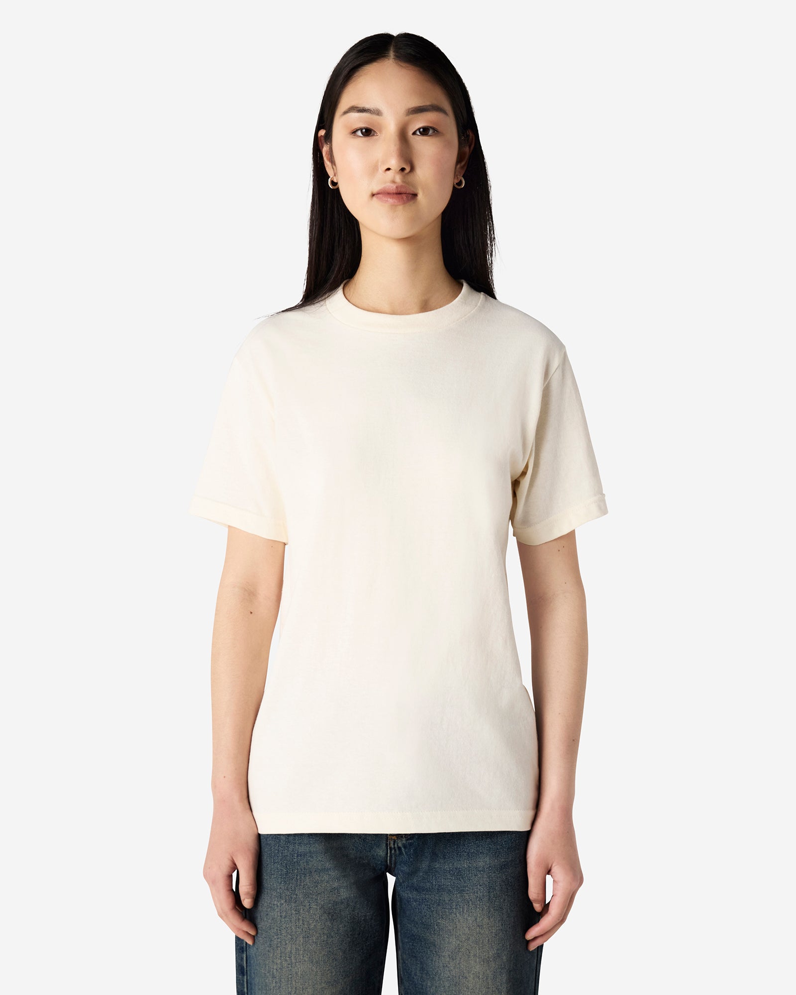 Female Model wearing a Cream Heavyweight Unisex Short Sleeve Crew Neck Tee, paired with blue jeans, standing against a plain background (front pose) -cream