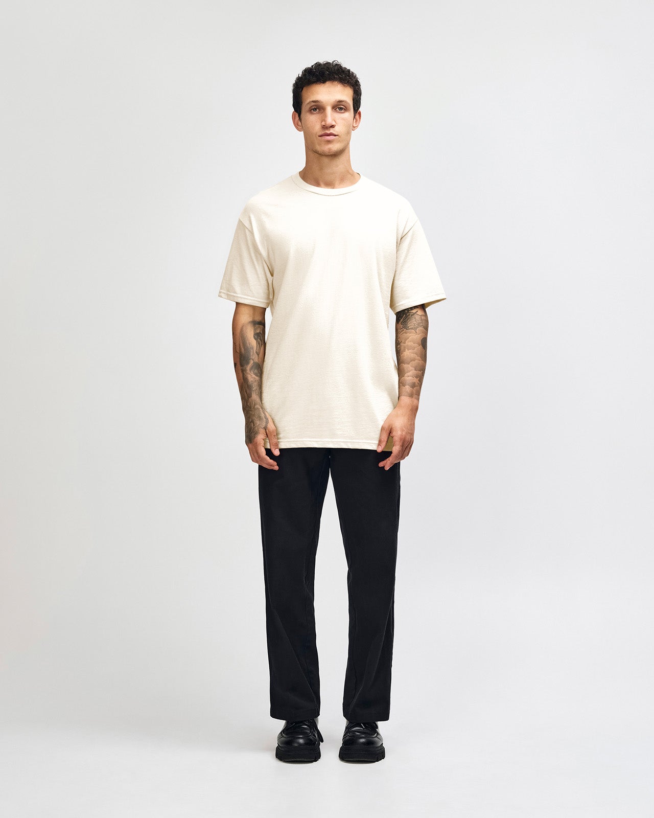 Male Model wearing a Charcoal Heavyweight Unisex Short Sleeve Crew Neck Tee, paired with blue jeans, standing against a plain background (front pose) -cream