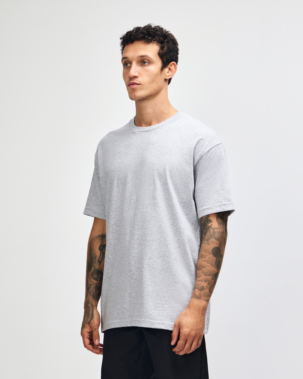 Male Model wearing a Heather Grey Heavyweight Unisex Short Sleeve Crew Neck Tee, paired with blue jeans, standing against a plain background (front pose) -heather grey