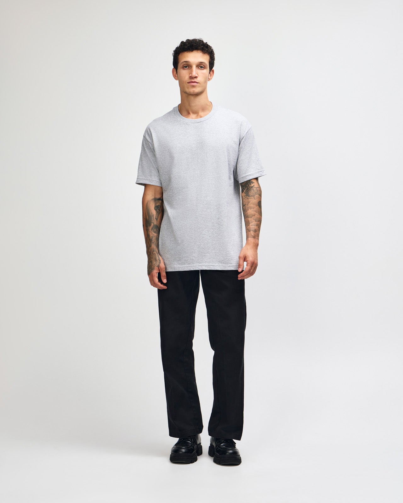 Male Model wearing a Heather Grey Heavyweight Unisex Short Sleeve Crew Neck Tee, paired with blue jeans, standing against a plain background (front pose) -heather grey