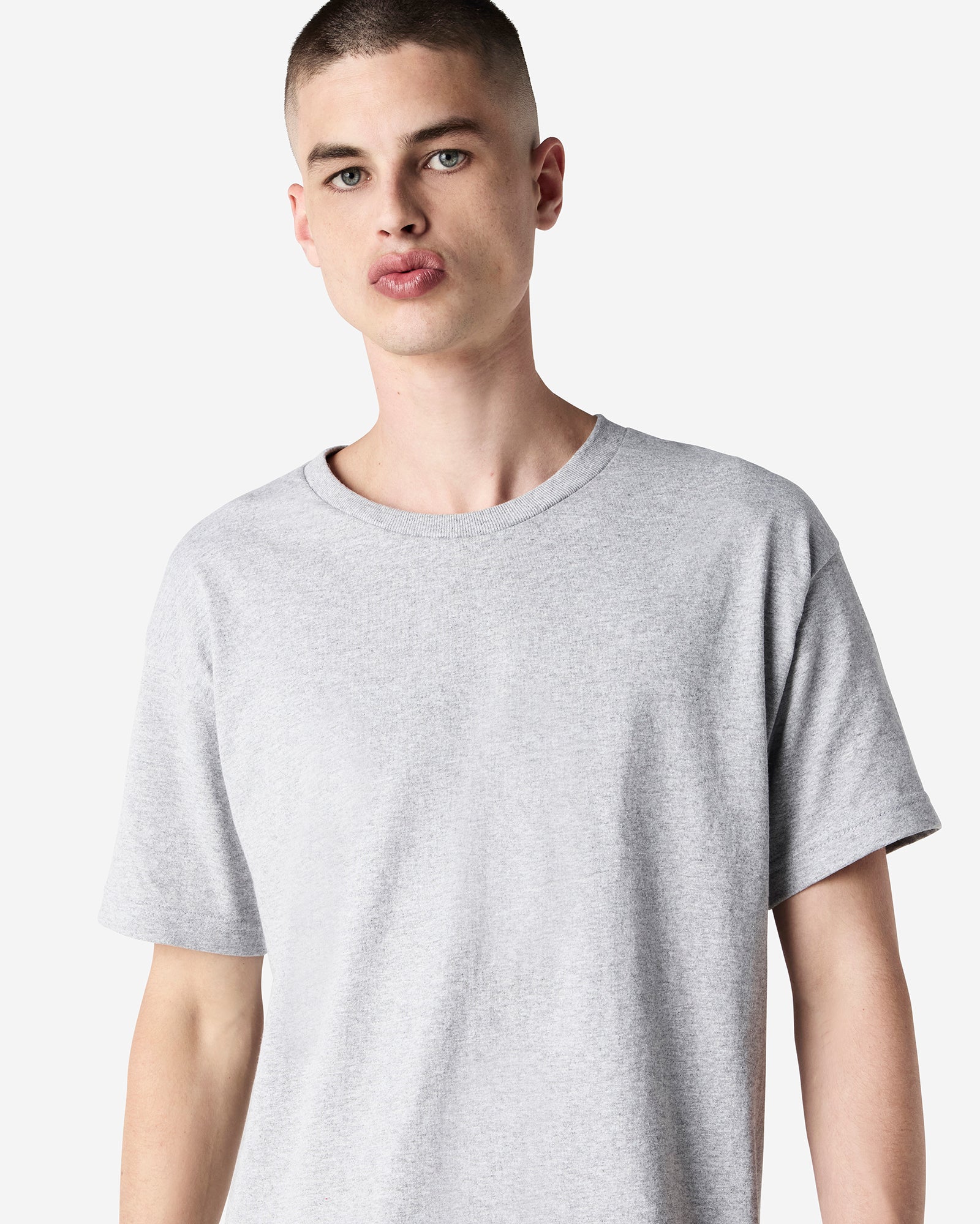 Male Model wearing a Heather Grey Heavyweight Unisex Short Sleeve Crew Neck Tee, paired with blue jeans, standing against a plain background (front pose) -heather grey
