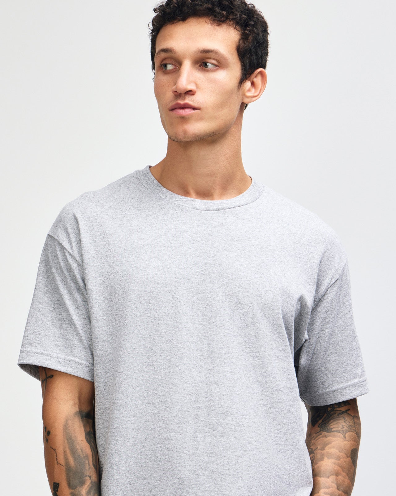 Male Model wearing a Heather Grey Heavyweight Unisex Short Sleeve Crew Neck Tee, paired with blue jeans, standing against a plain background (front pose) -heather grey