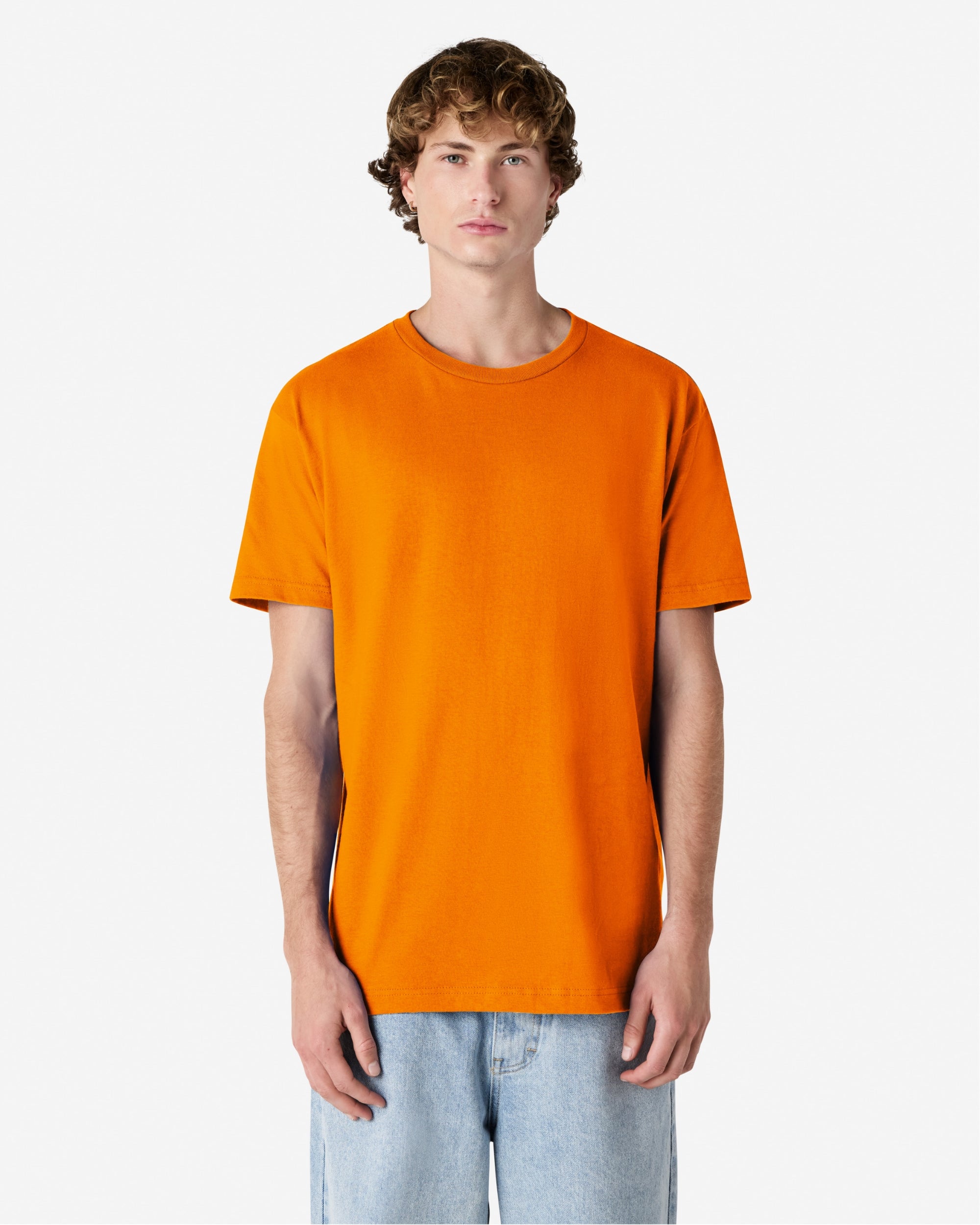 Male Model wearing a orange Heavyweight Unisex Short Sleeve Crew Neck Tee, paired with blue jeans, standing against a plain background (front pose) -orange