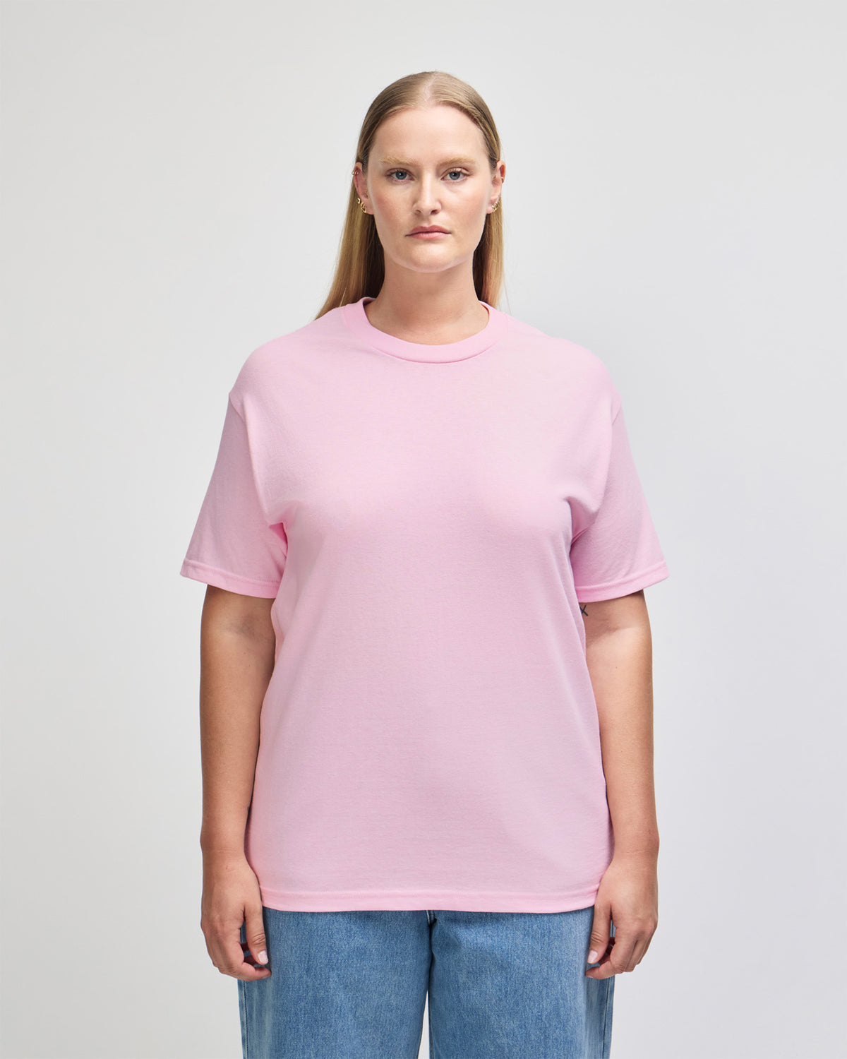 Female Model wearing a Pink Heavyweight Unisex Short Sleeve Crew Neck Tee, paired with blue jeans, standing against a plain background (front pose) - pink