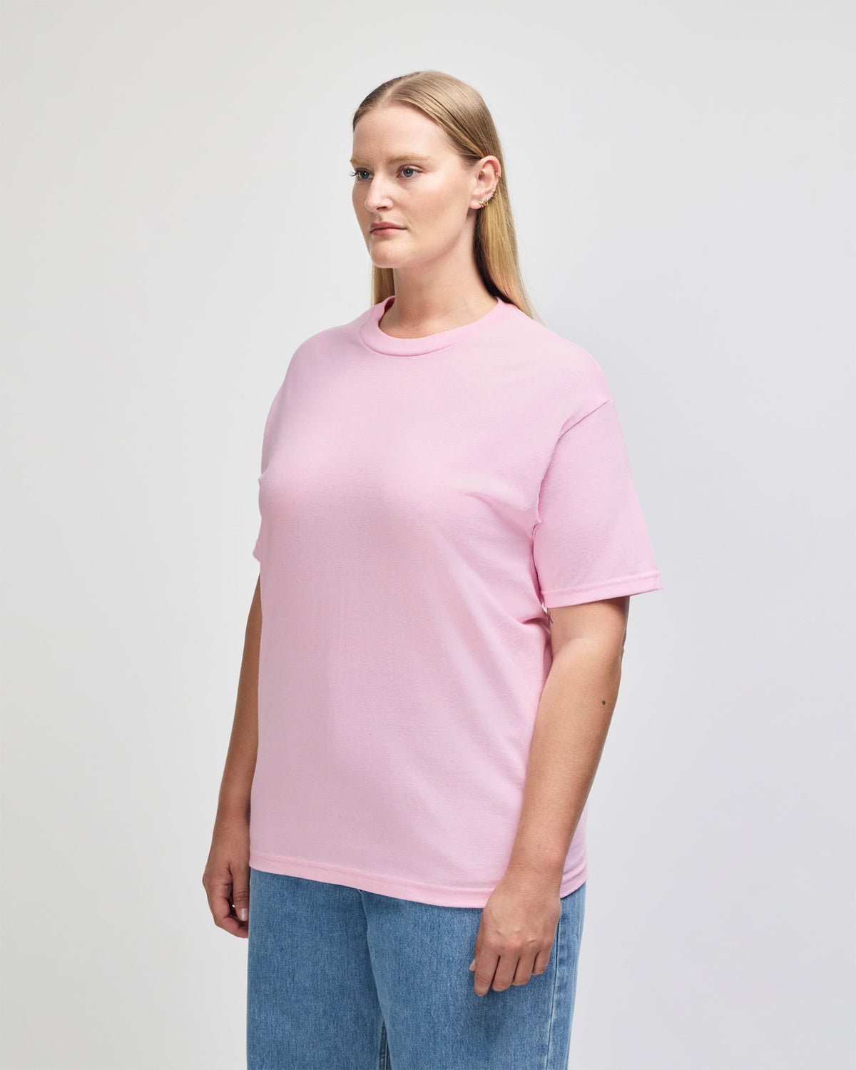 Female Model wearing a Pink Heavyweight Unisex Short Sleeve Crew Neck Tee, paired with blue jeans, standing against a plain background (side pose) - pink