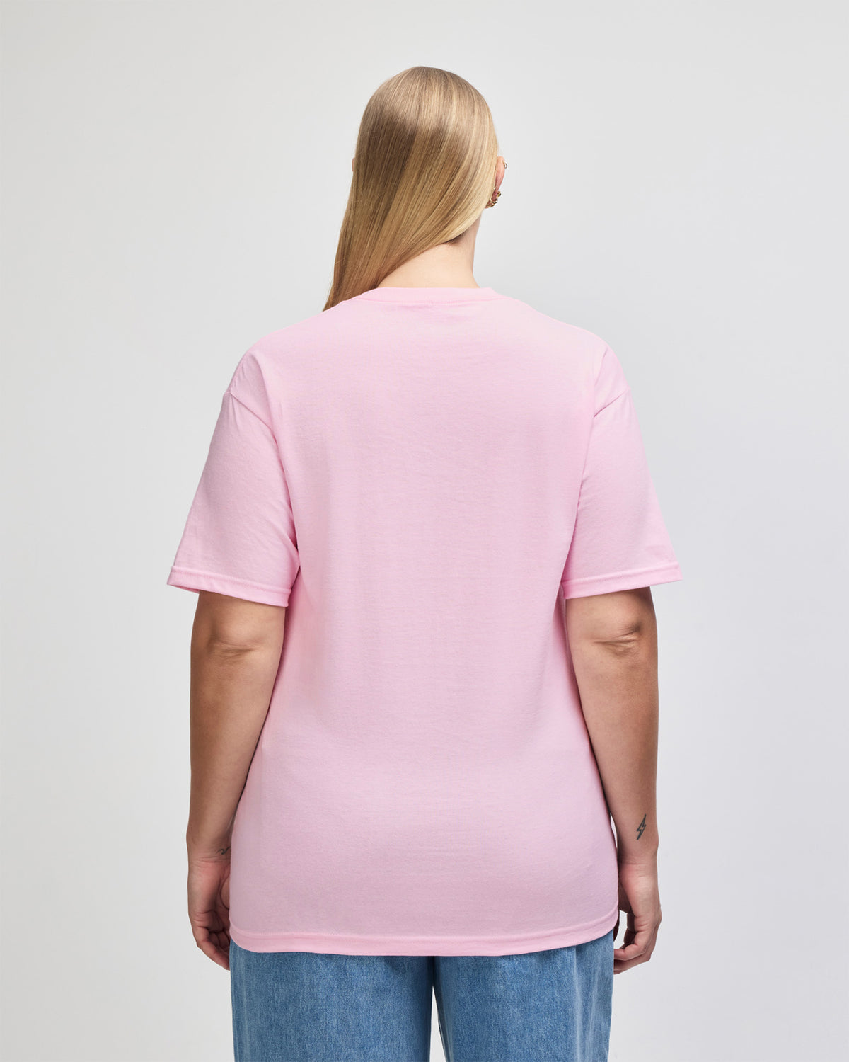 Female Model wearing a Pink Heavyweight Unisex Short Sleeve Crew Neck Tee, paired with blue jeans, standing against a plain background (back pose) - pink