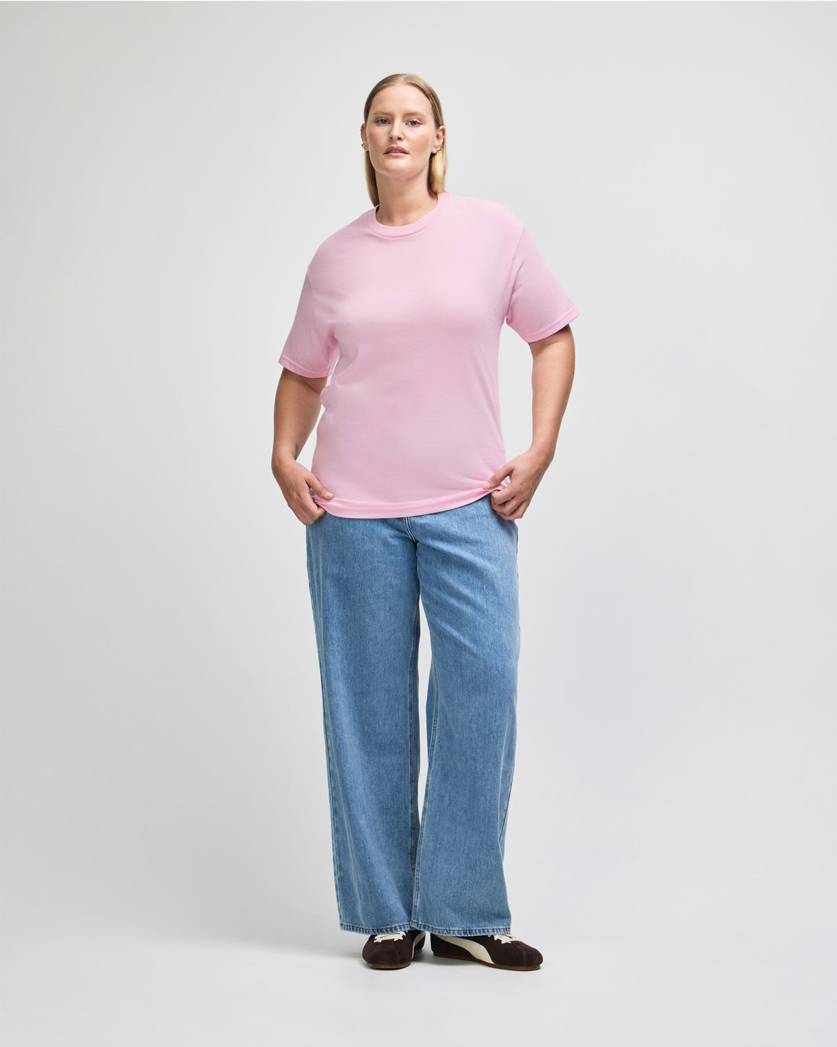 Female Model wearing a Pink Heavyweight Unisex Short Sleeve Crew Neck Tee, paired with blue jeans, standing against a plain background (front pose) - pink