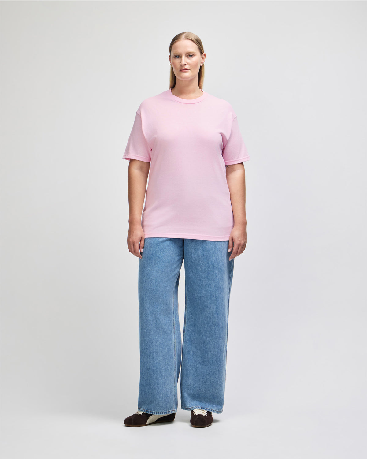 Female Model wearing a Pink Heavyweight Unisex Short Sleeve Crew Neck Tee, paired with blue jeans, standing against a plain background (front pose) - pink
