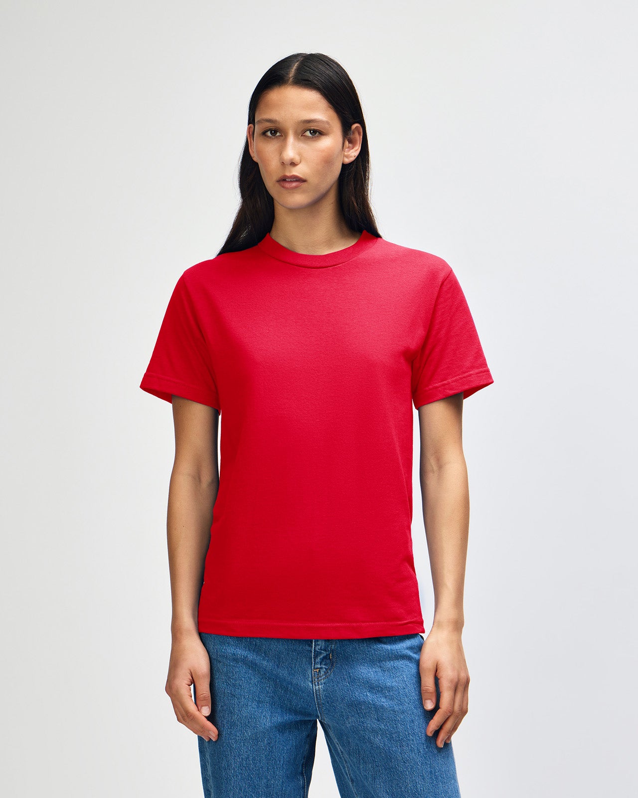 Female Model wearing a red Heavyweight Unisex Short Sleeve Crew Neck Tee, paired with blue jeans, standing against a plain background (front pose) -red