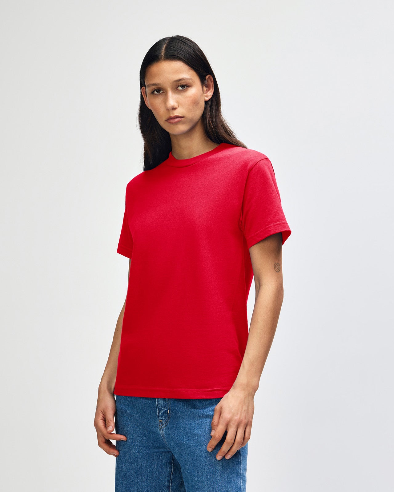 Female Model wearing a red Heavyweight Unisex Short Sleeve Crew Neck Tee, paired with blue jeans, standing against a plain background (front pose) -red