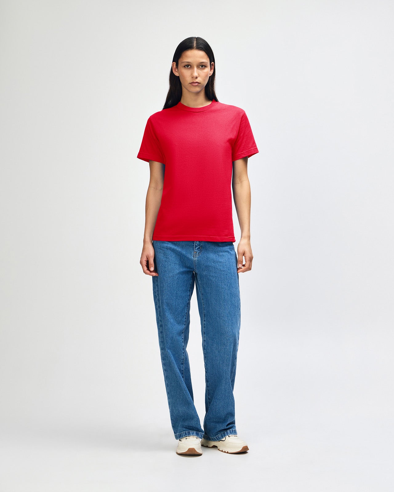 Female Model wearing a red Heavyweight Unisex Short Sleeve Crew Neck Tee, paired with blue jeans, standing against a plain background (front pose) -red
