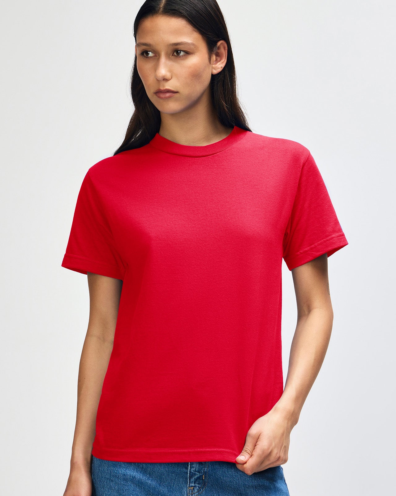 Female Model wearing a red Heavyweight Unisex Short Sleeve Crew Neck Tee, paired with blue jeans, standing against a plain background (front pose) -red