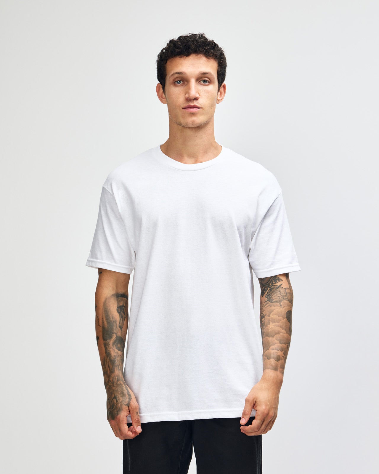Male Model wearing a white Heavyweight Unisex Short Sleeve Crew Neck Tee, paired with blue jeans, standing against a plain background (front pose) -white