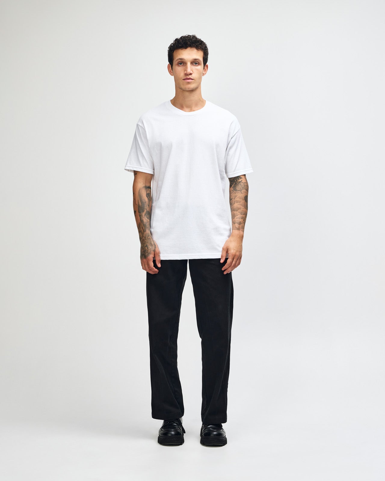 Male Model wearing a white Heavyweight Unisex Short Sleeve Crew Neck Tee, paired with blue jeans, standing against a plain background (front pose) -white