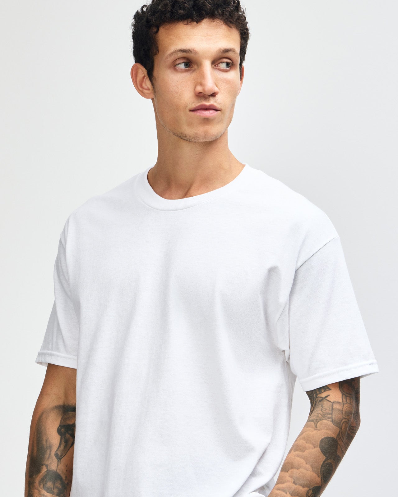 Male Model wearing a white Heavyweight Unisex Short Sleeve Crew Neck Tee, paired with blue jeans, standing against a plain background (front pose) -white