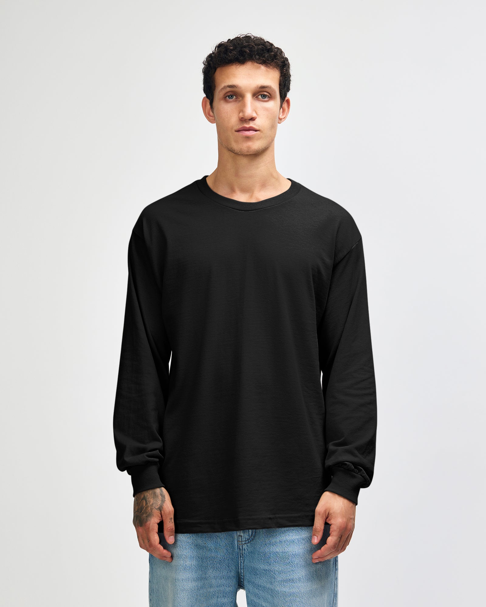 front view of a male Model wearing a black coloured Heavyweight Unisex Long Sleeve Crew Neck Tee, paired with blue jeans, standing against a plain background (front pose) -black