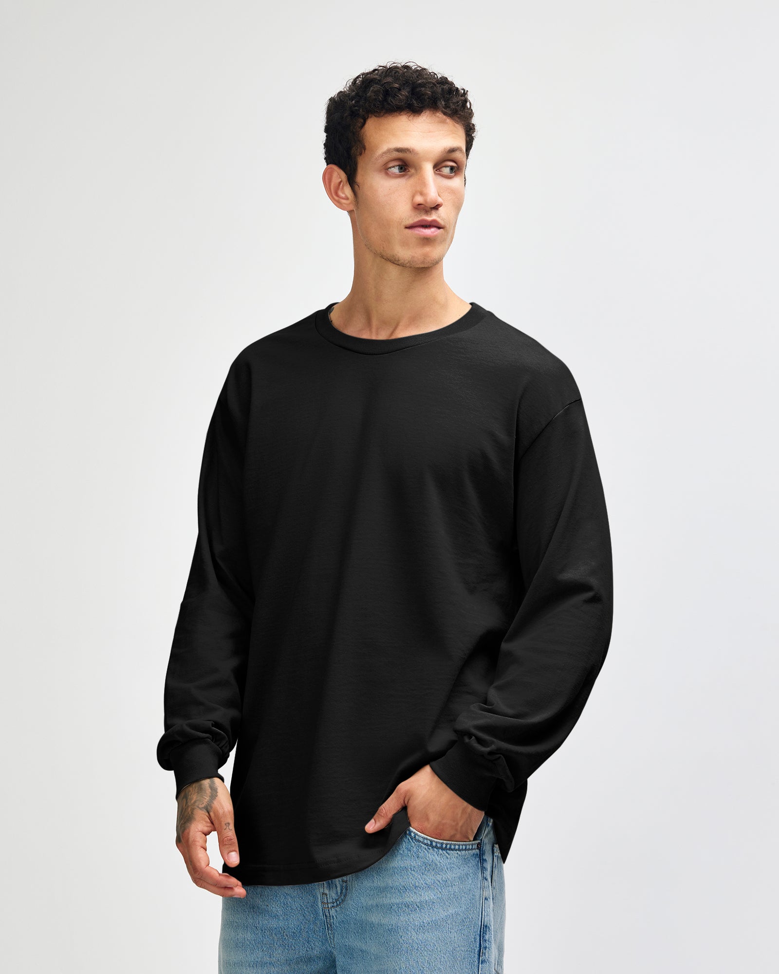 front view of a male Model wearing a black coloured Heavyweight Unisex Long Sleeve Crew Neck Tee, paired with blue jeans, standing against a plain background (front pose) -black