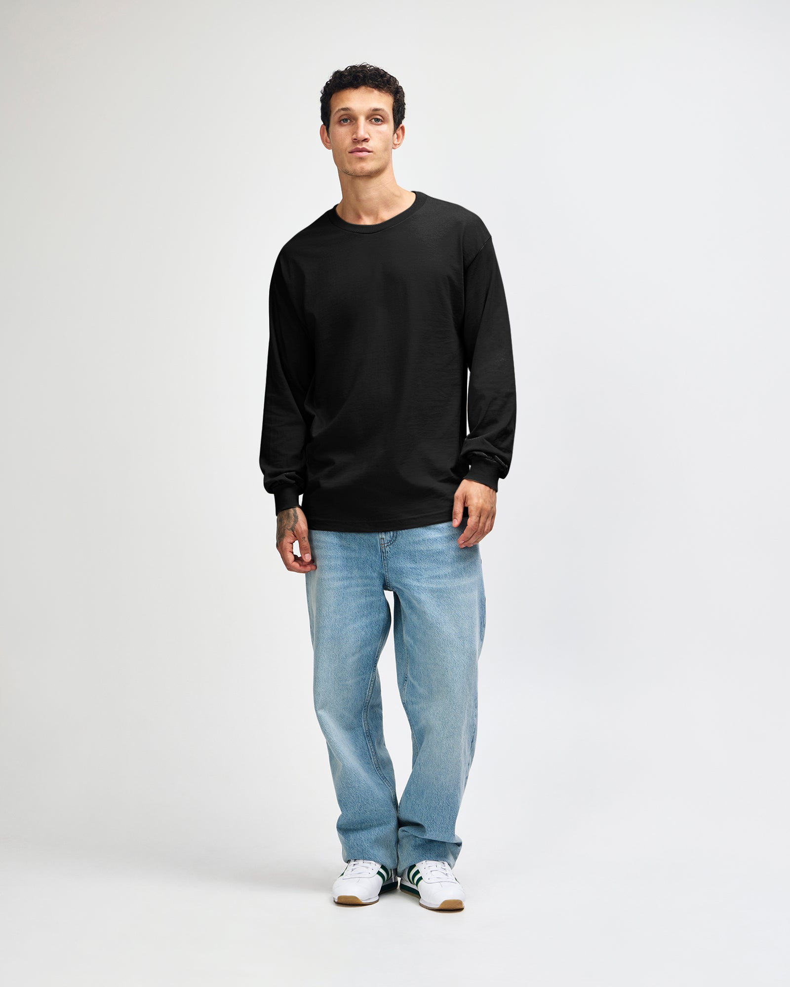 front view of a male Model wearing a black coloured Heavyweight Unisex Long Sleeve Crew Neck Tee, paired with blue jeans, standing against a plain background (front pose) -black