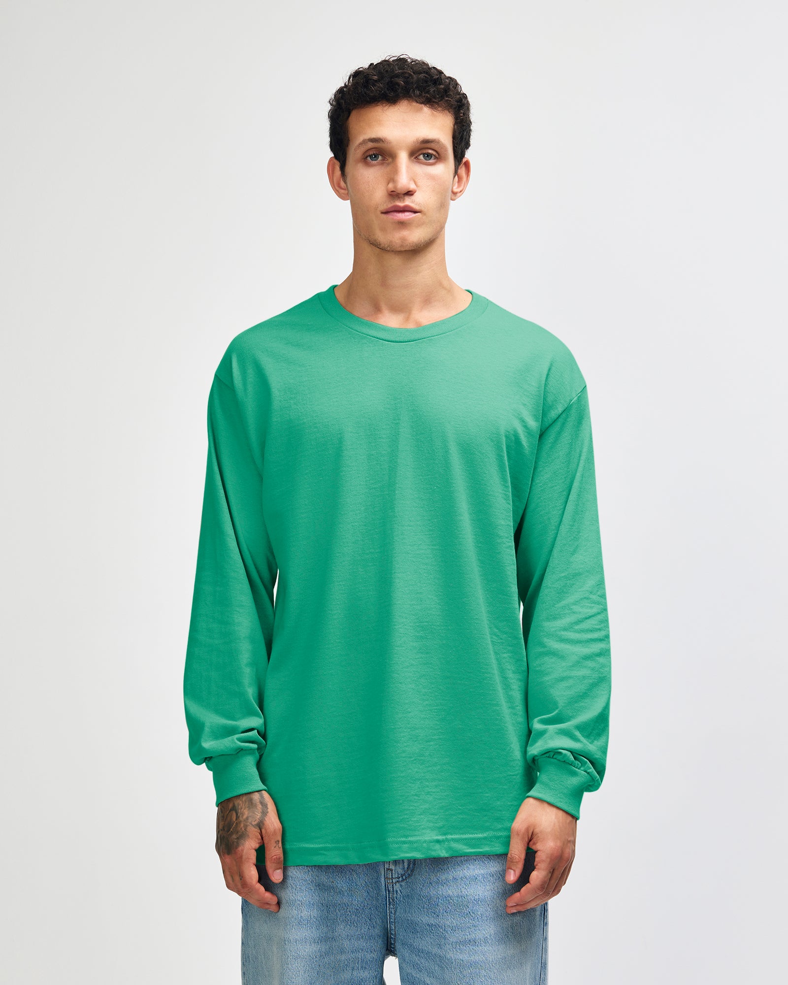 front view of a male Model wearing a Celadon colored Heavyweight Unisex Long Sleeve Crew Neck Tee, paired with blue jeans, standing against a plain background (front pose) -celadon