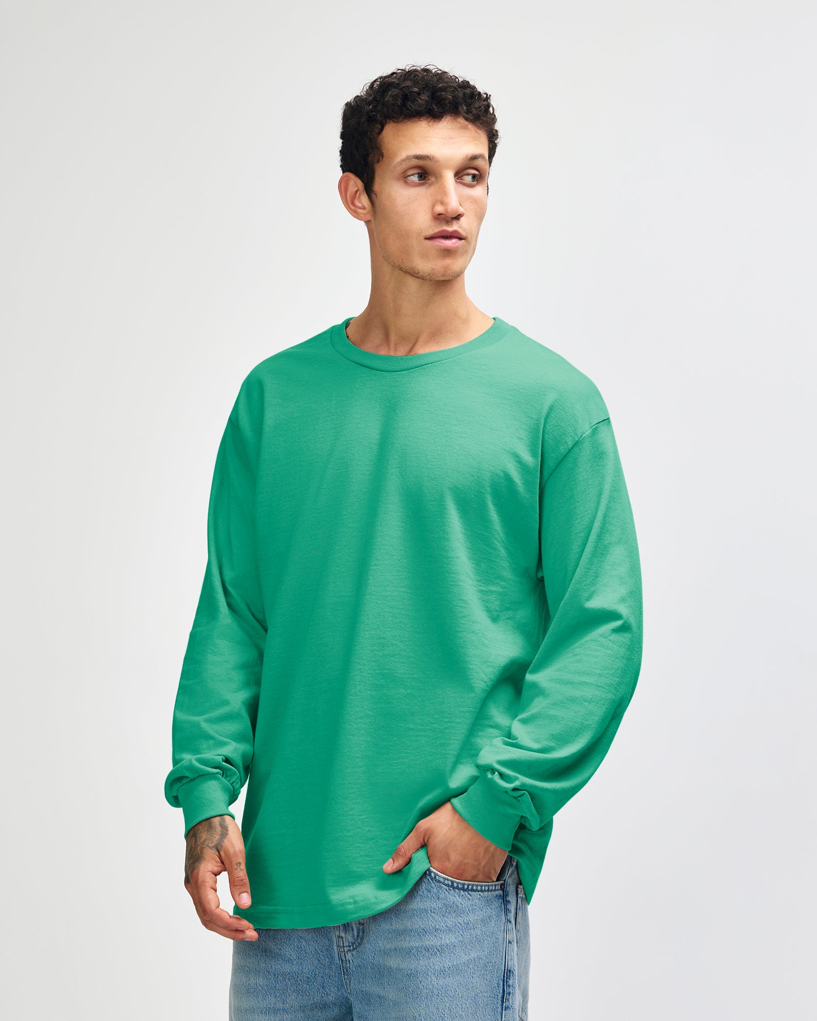 front view of a male Model wearing a Celadon colored Heavyweight Unisex Long Sleeve Crew Neck Tee, paired with blue jeans, standing against a plain background (front pose) -celadon