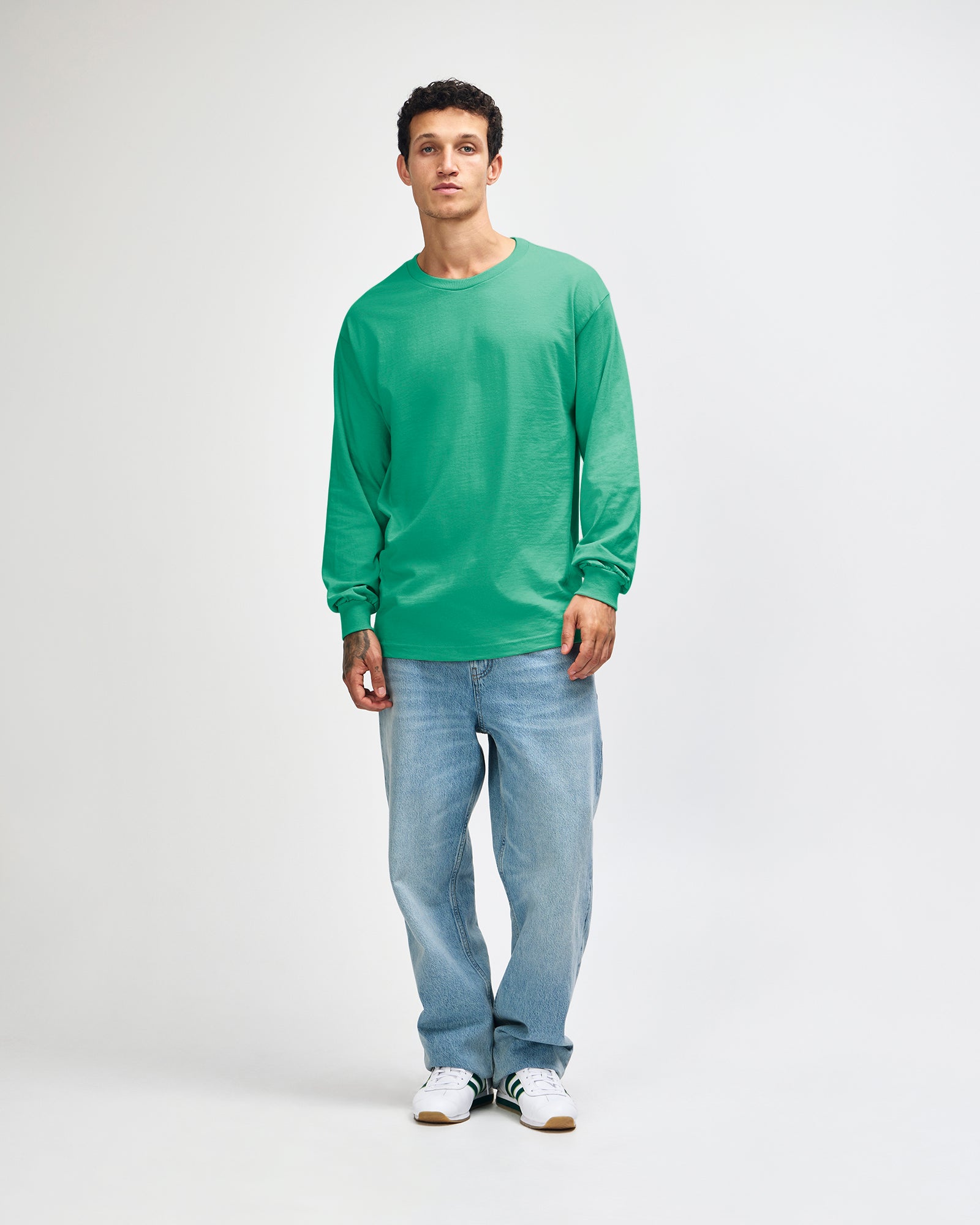 front view of a male Model wearing a Celadon colored Heavyweight Unisex Long Sleeve Crew Neck Tee, paired with blue jeans, standing against a plain background (front pose) -celadon