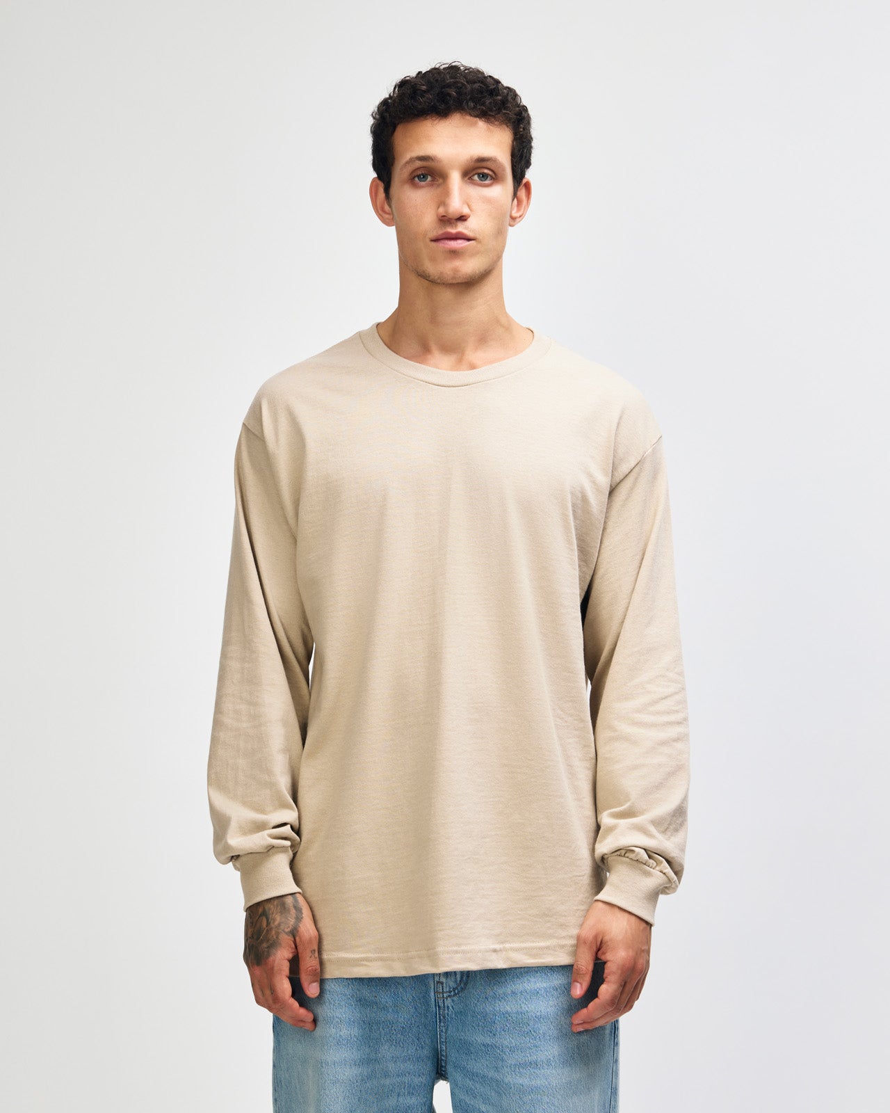 front view of a male Model wearing a Sand colored Heavyweight Unisex Long Sleeve Crew Neck Tee, paired with blue jeans, standing against a plain background (front pose) -sand