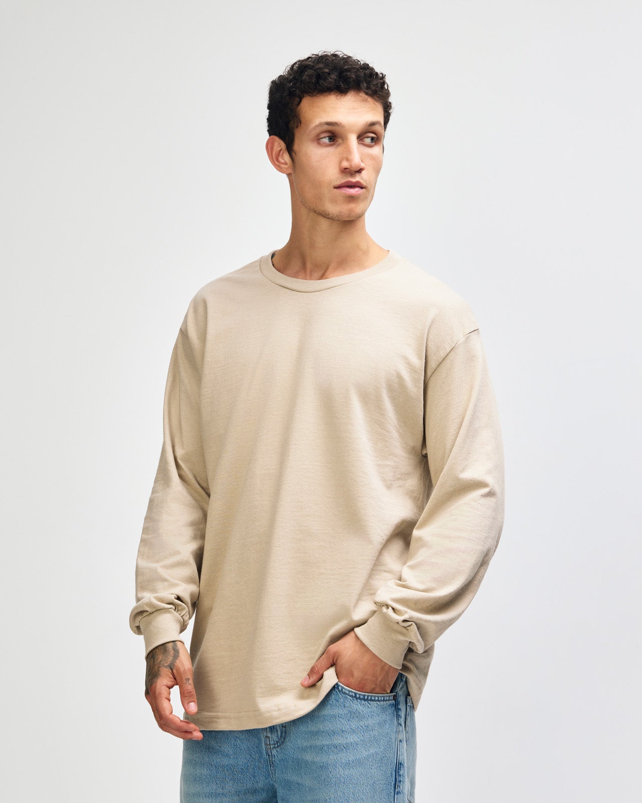 front view of a male Model wearing a Sand colored Heavyweight Unisex Long Sleeve Crew Neck Tee, paired with blue jeans, standing against a plain background (front pose) -sand