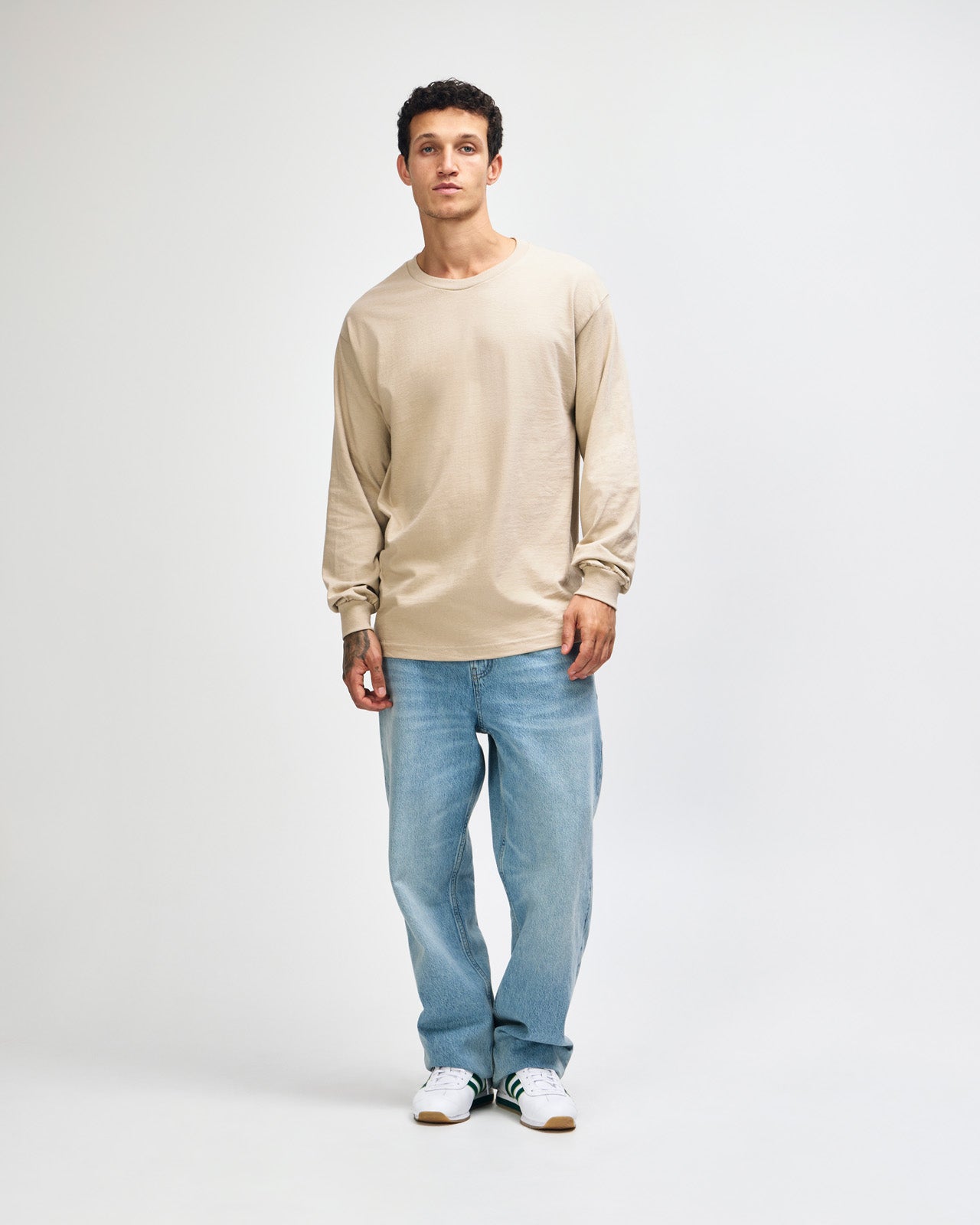 front view of a male Model wearing a Sand colored Heavyweight Unisex Long Sleeve Crew Neck Tee, paired with blue jeans, standing against a plain background (front pose) -sand