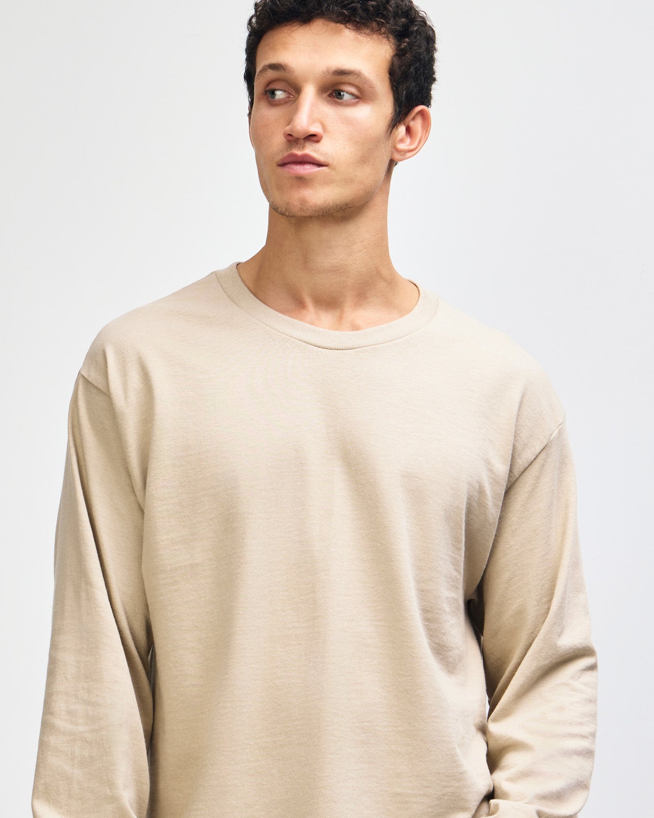 front view of a male Model wearing a Sand colored Heavyweight Unisex Long Sleeve Crew Neck Tee, paired with blue jeans, standing against a plain background (front pose) -sand