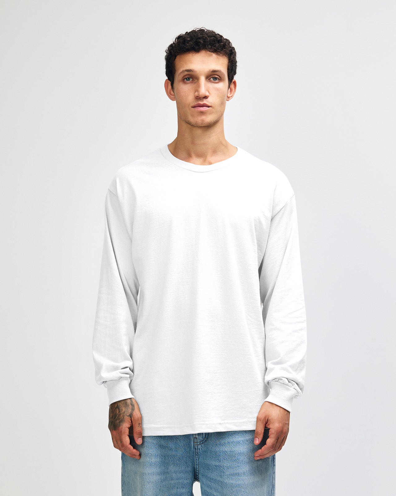 front view of a male Model wearing a White colored Heavyweight Unisex Long Sleeve Crew Neck Tee, paired with blue jeans, standing against a plain background (front pose) -white
