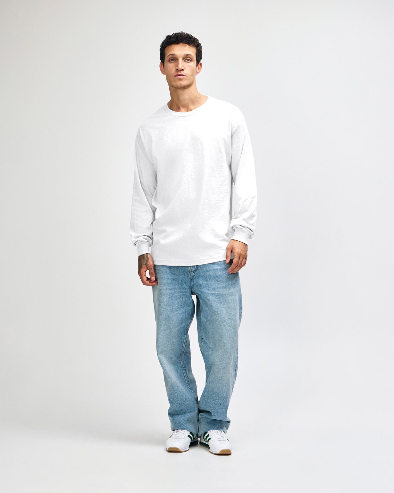 front view of a male Model wearing a White colored Heavyweight Unisex Long Sleeve Crew Neck Tee, paired with blue jeans, standing against a plain background (front pose) -white