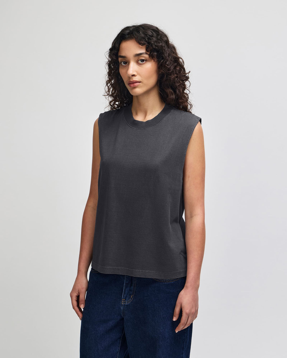 A woman with curly dark hair wearing a dark charcoal gray sleeveless crop top and dark blue jeans, standing against a light gray studio background. -Faded Black