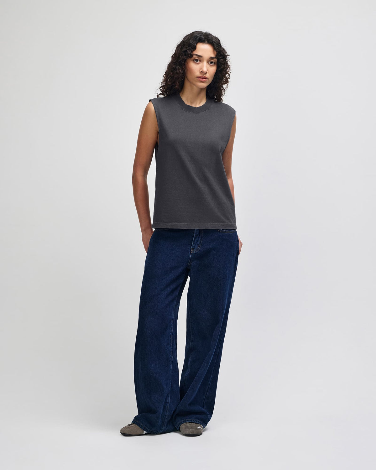 A model stands against a light gray studio background wearing a dark charcoal gray sleeveless top with shoulder tab details and dark blue wide-leg jeans. -Faded Black