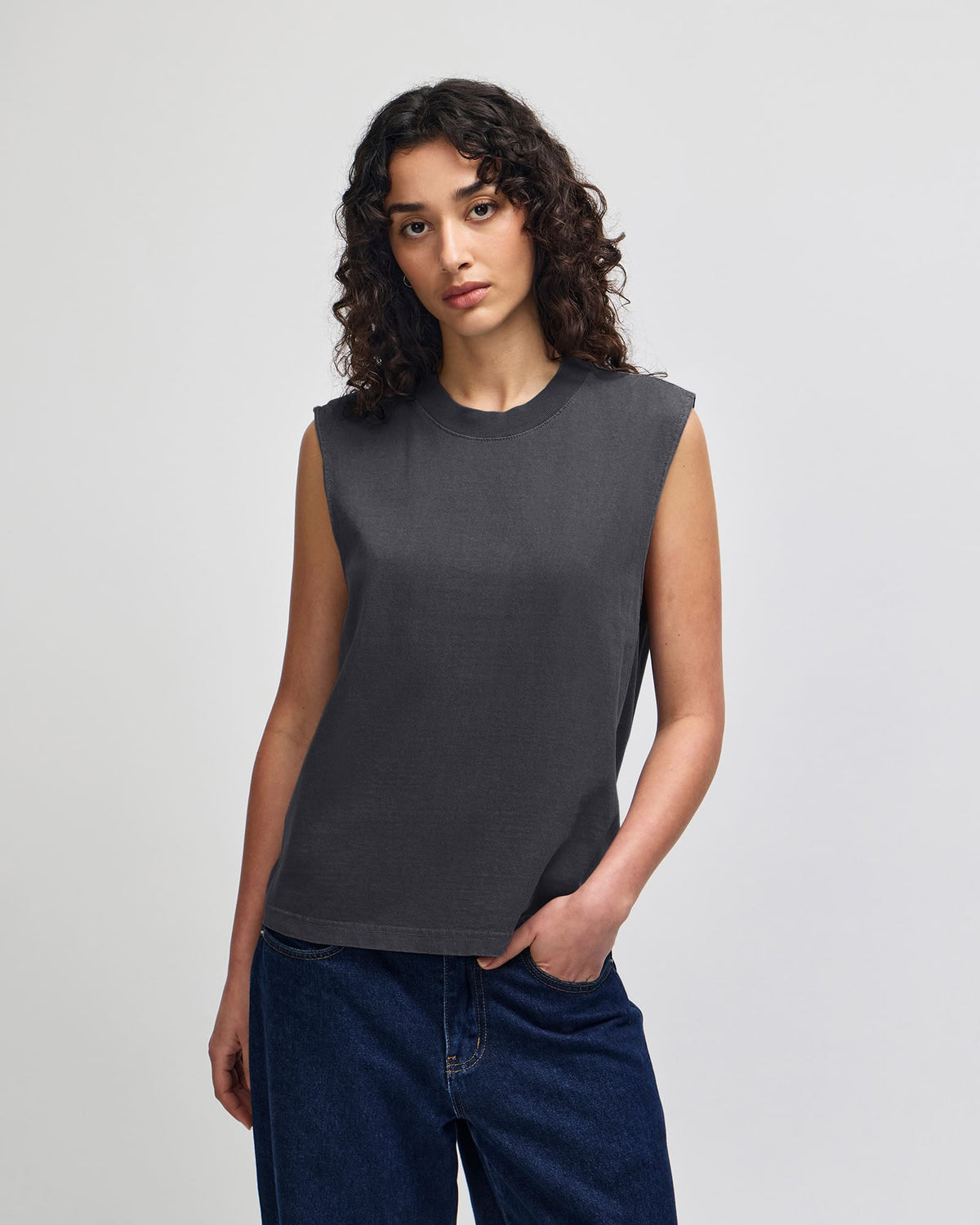 A woman with curly dark hair wearing a dark charcoal gray sleeveless crewneck top and dark blue jeans, standing against a light gray studio background. -Faded Black