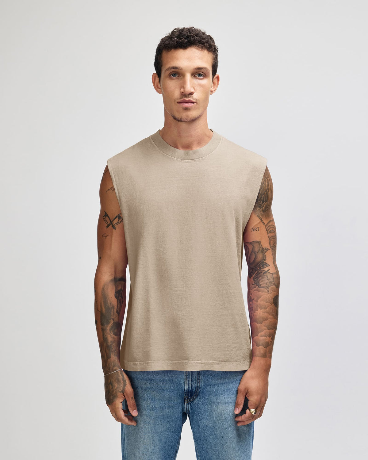 A man wearing a sleeveless beige t-shirt and blue jeans, standing against a light gray studio background. -Faded Bone