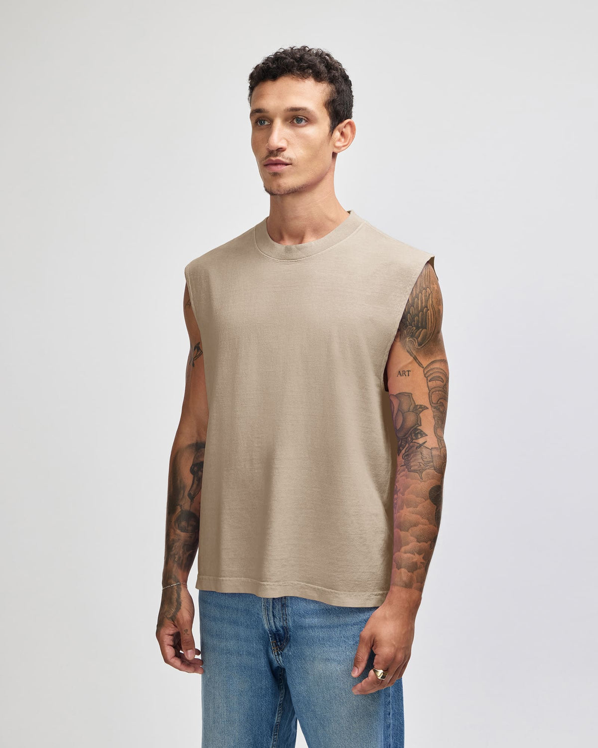 A man wearing a sleeveless beige crewneck t-shirt and blue jeans, standing against a light gray studio background. -Faded Bone