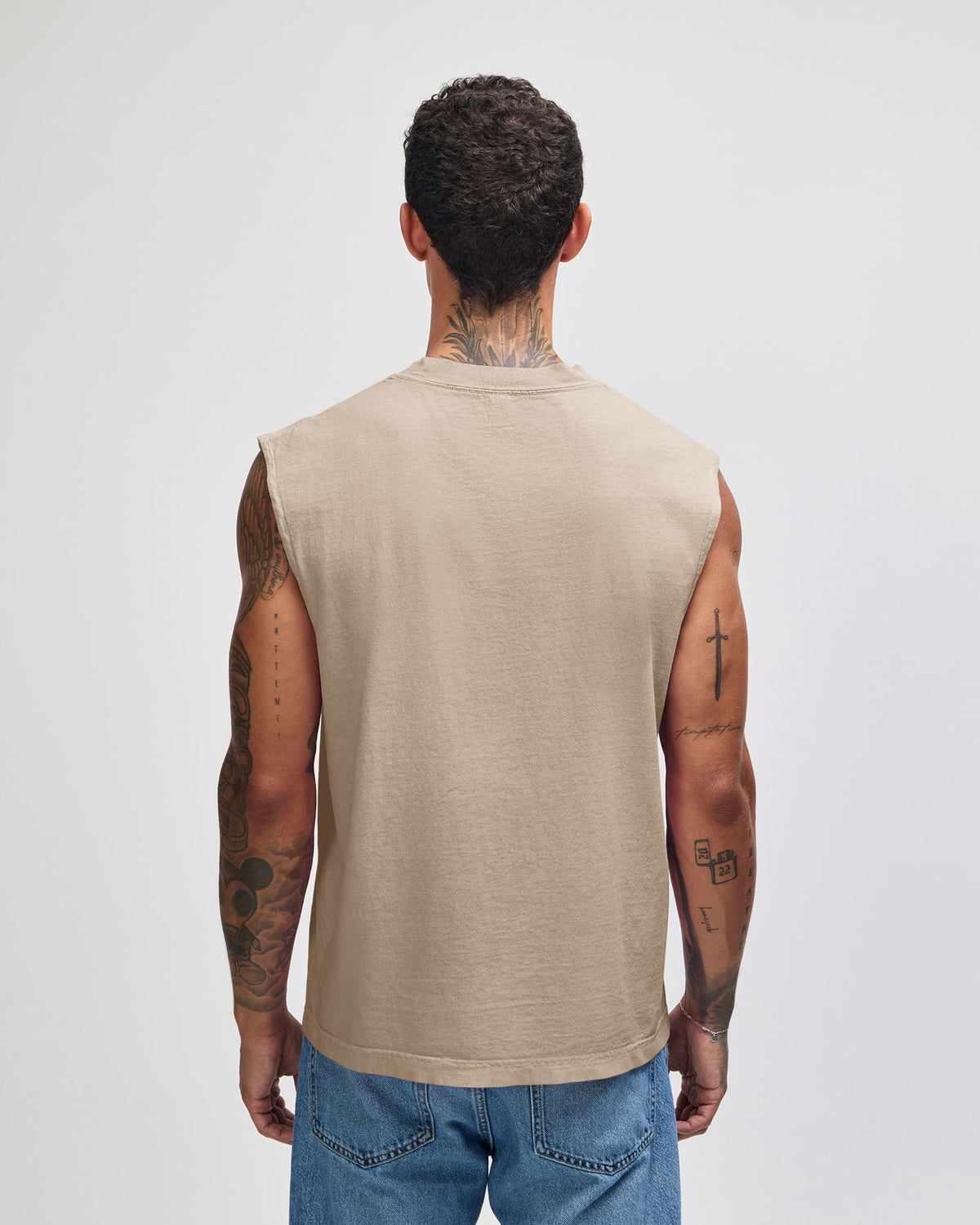 A man viewed from behind wearing a sleeveless beige t-shirt and blue jeans, standing against a light gray studio background -Faded Bone