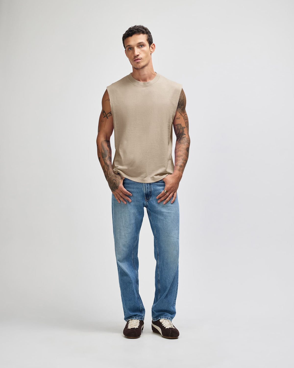 A man modeling a beige sleeveless crewneck shirt and blue jeans against a light gray studio background -Faded Bone