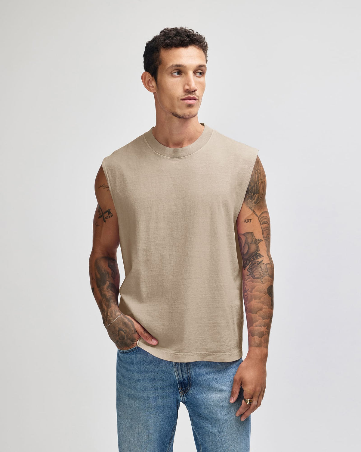 A man wearing a sleeveless beige linen-blend t-shirt and blue jeans, standing against a light gray studio background -Faded Bone