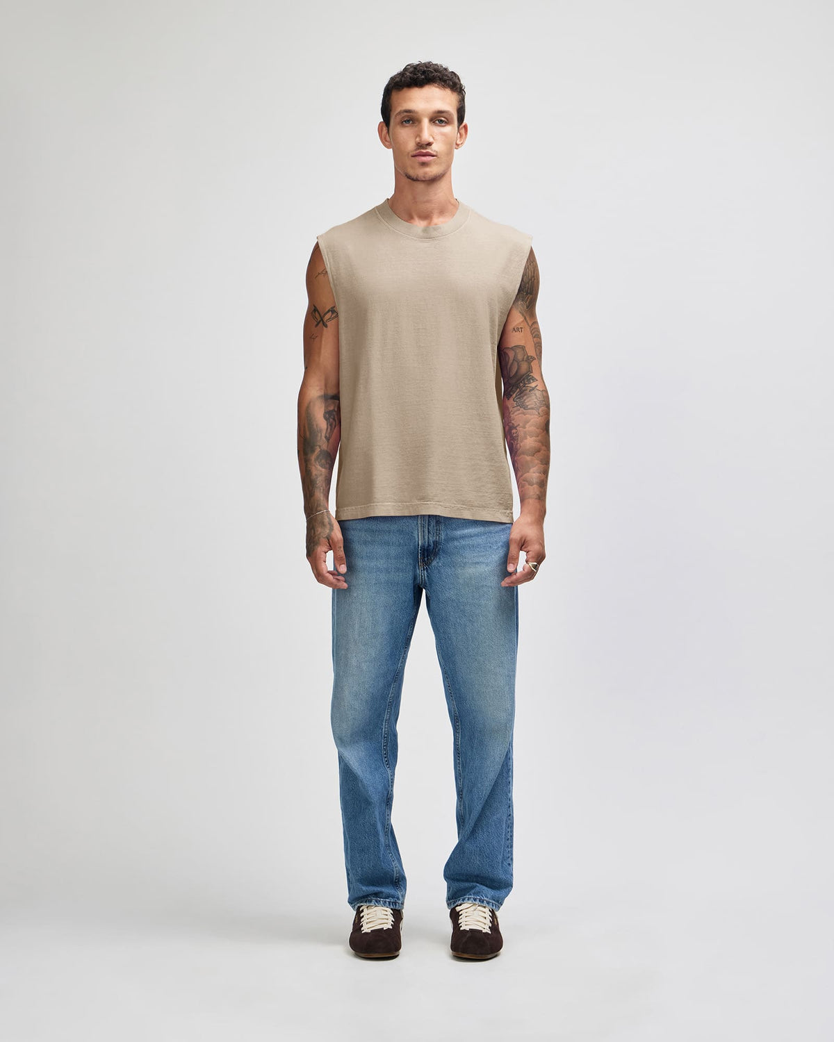 A man modeling a sleeveless beige t-shirt and blue jeans against a light gray studio background -Faded Bone