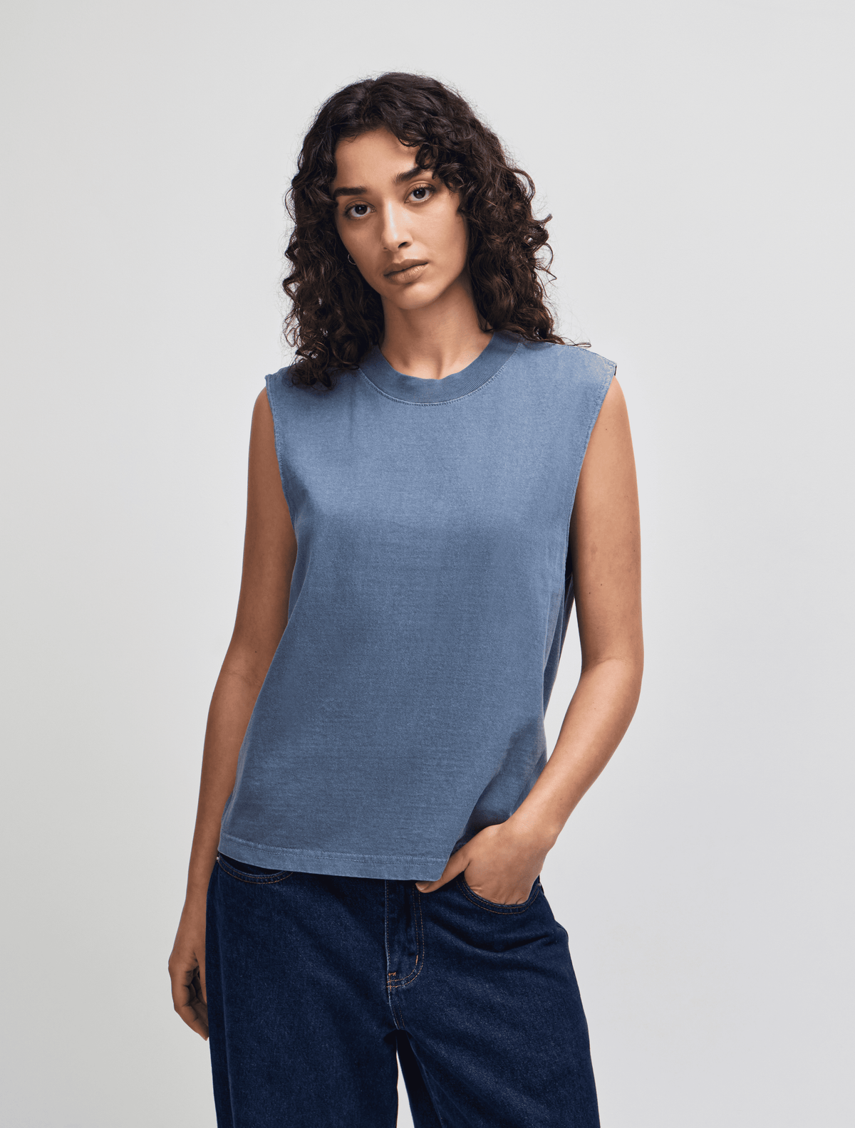 A woman modeling a sleeveless crewneck top in a dusty slate blue color, paired with dark blue jeans, against a plain light gray studio background. -Faded Navy