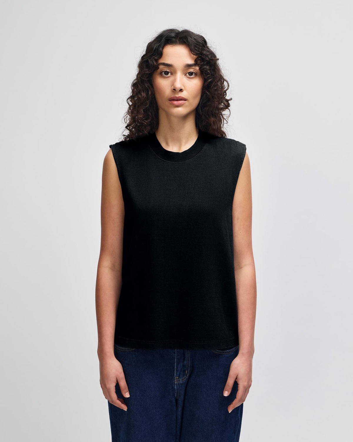 A woman with curly dark hair wearing a sleeveless black knit top and dark blue jeans, standing against a light gray studio background. -True Black