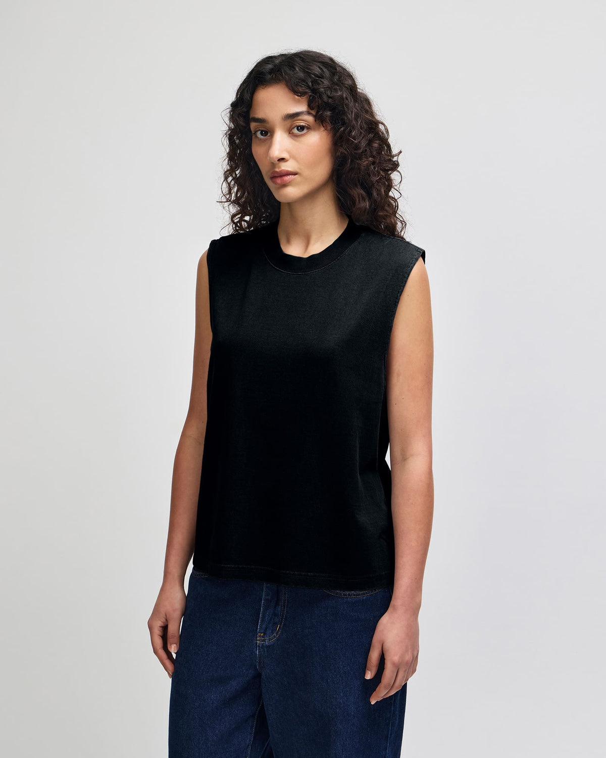 A woman with curly dark hair wearing a black sleeveless crewneck top and dark blue jeans, standing against a light gray studio background. -True Black