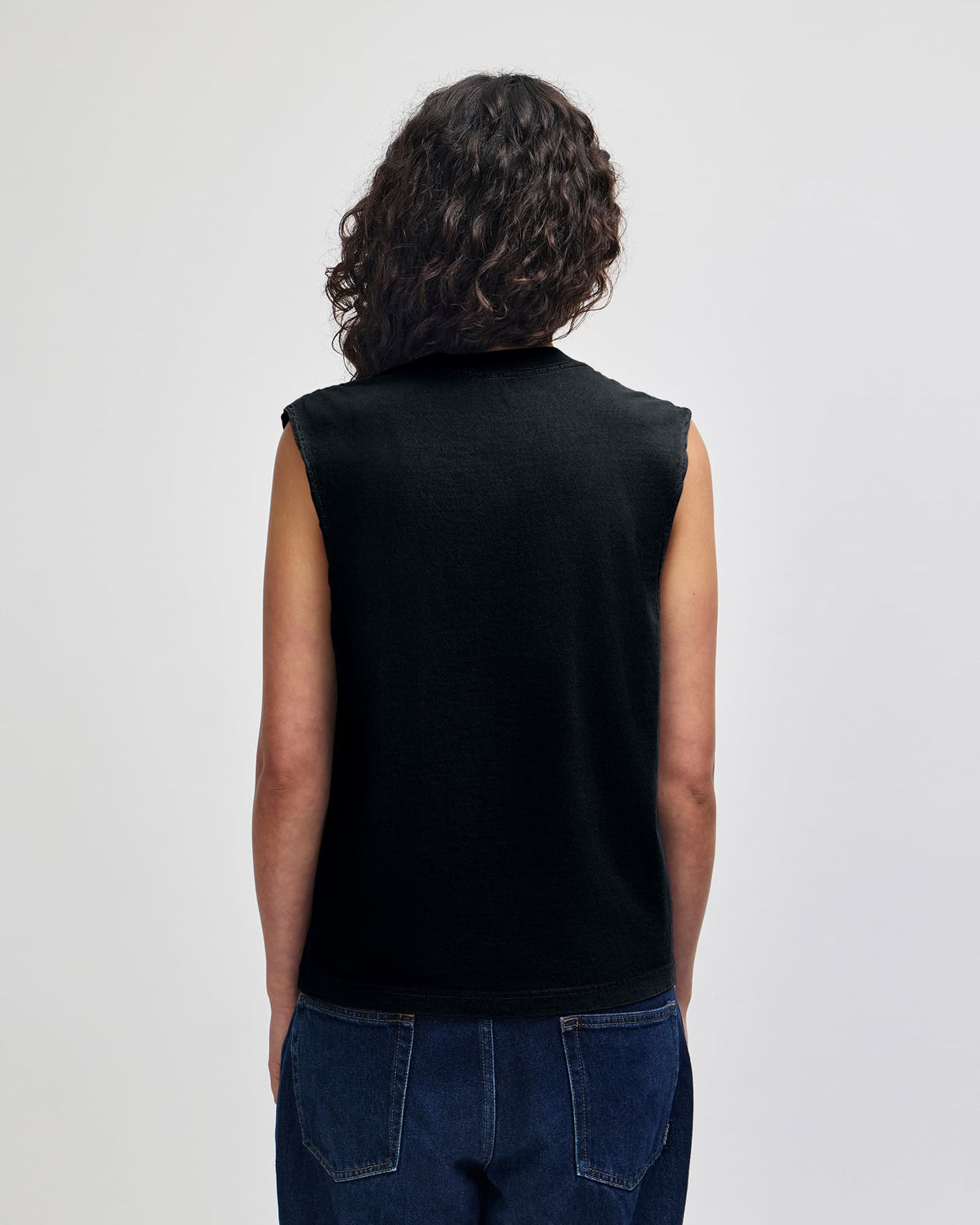 A person shown from the back wearing a black sleeveless knit top and dark blue jeans against a plain light gray studio background. -True Black