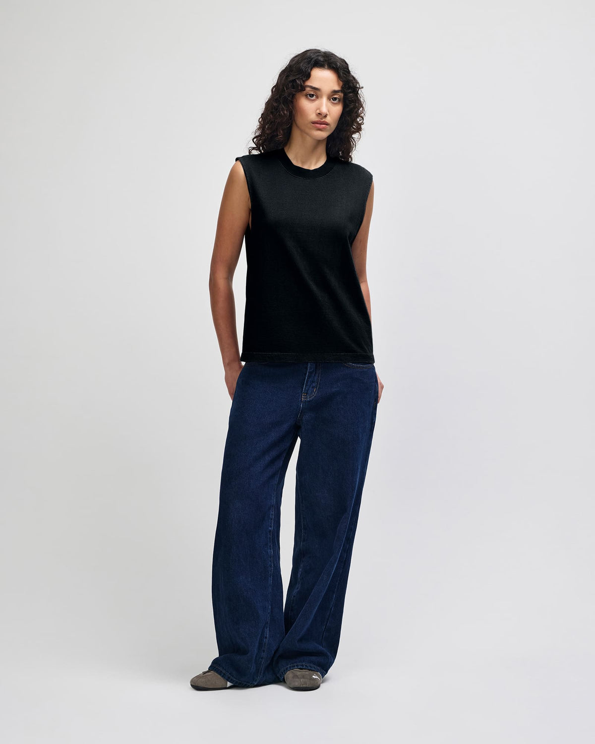 A model stands against a light gray studio background wearing a sleeveless black top and dark blue wide-leg jeans. -True Black