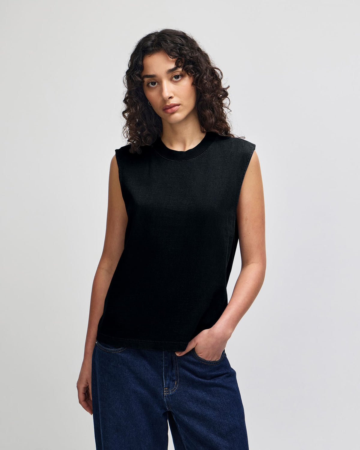 A woman modeling a sleeveless black knit top with a crew neck, paired with dark blue jeans, against a light gray studio background. -True Black